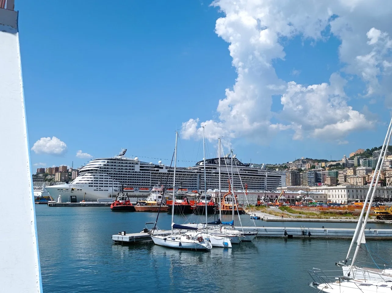 Navio MSC Meraviglia no porto de Gênova, com veleiros em primeiro plano e céu azul.