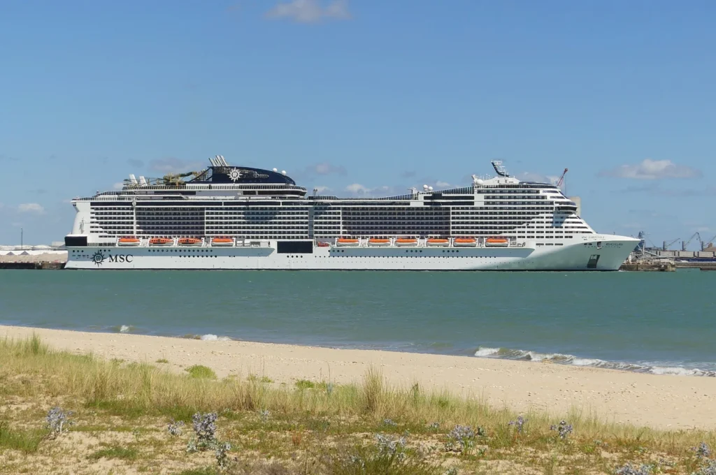 Navio MSC Virtuosa visto de perfil no porto de La Rochelle, com água e faixa de areia em primeiro plano.