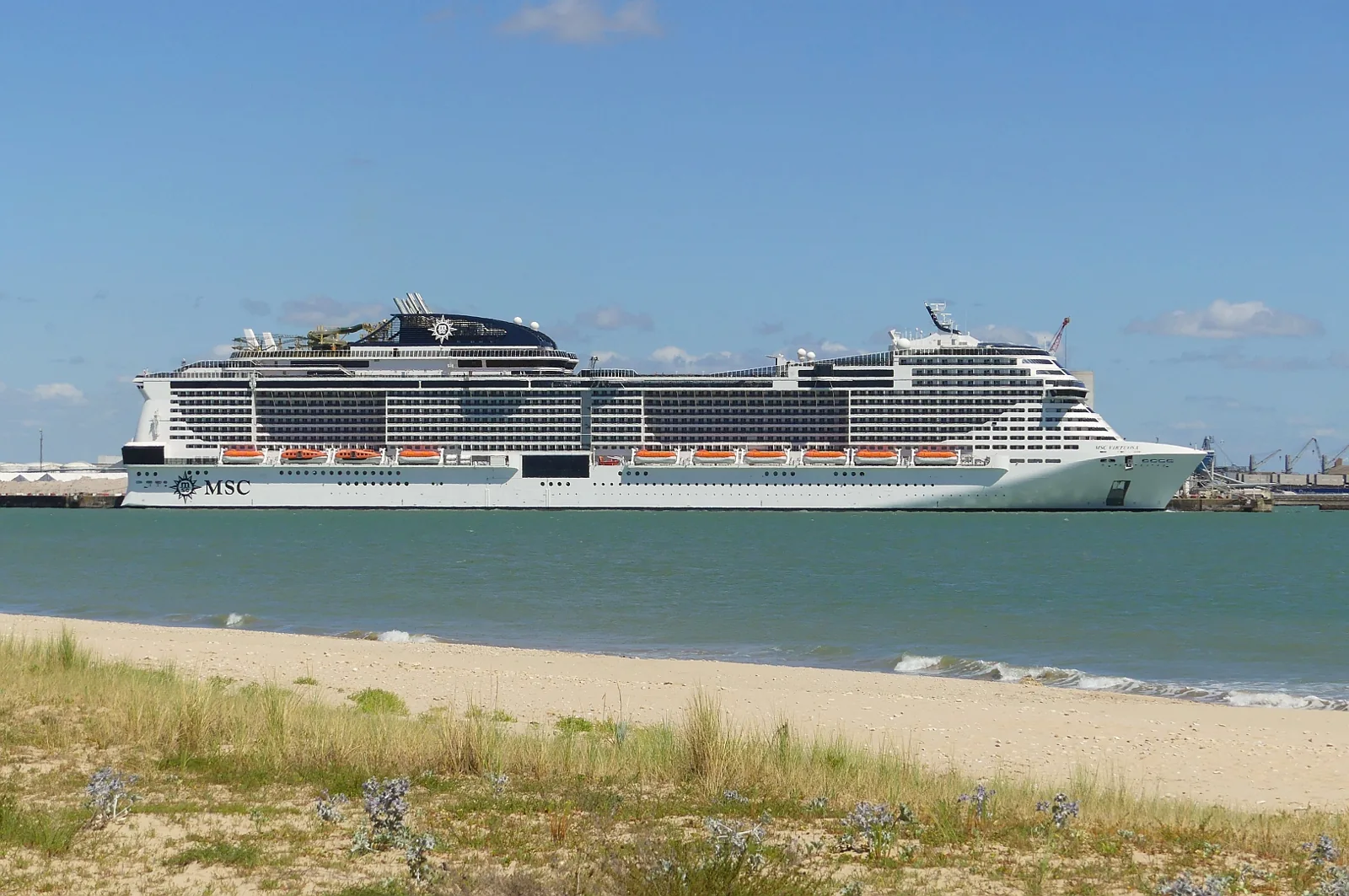 Navio MSC Virtuosa visto de perfil no porto de La Rochelle, com água e faixa de areia em primeiro plano.