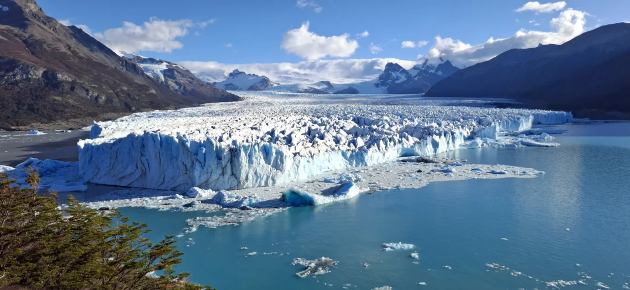 Vista panorâmica do glaciar Perito Moreno, com paredes de gelo, blocos flutuantes no lago e montanhas ao fundo.