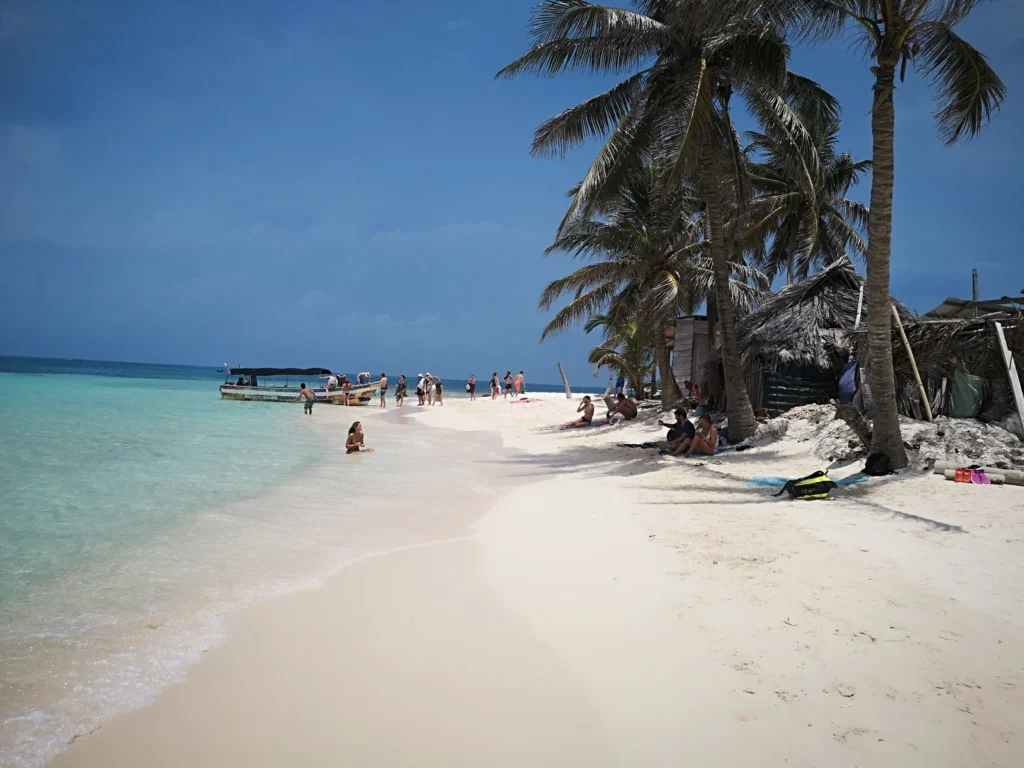 Praia de San Blas com areia clara, coqueiros e visitantes junto a uma lancha no mar.