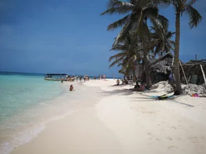 Praia de San Blas com areia clara, coqueiros e visitantes junto a uma lancha no mar.