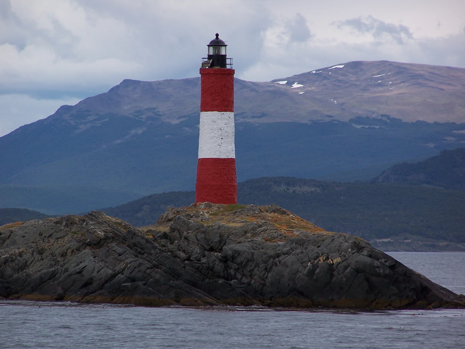 Farol Les Éclaireurs, vermelho e branco, sobre uma ilhota no Canal de Beagle, com montanhas ao fundo.